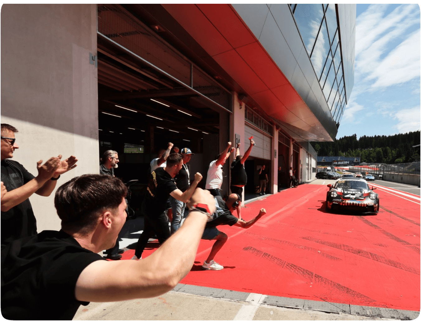 Zespół Fabiana Dybionki eksploduje radością przy boksie na torze Red Bull Ring w Austrii, witając go po zwycięskim przejeździe. Fabian w swoim Porsche 911 GT3 Cup z numerem 64 i grafiką rekina zjeżdża do alei serwisowej po wygraniu swojego pierwszego wyścigu w serii Porsche Deutschland Cup.