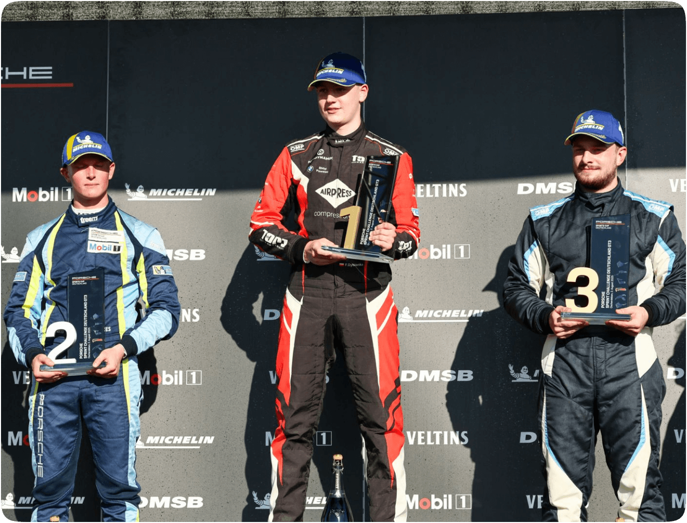 Fabian Dybionka stoi na najwyższym stopniu podium z trofeum za pierwsze miejsce po wyścigu serii Porsche Deutschland Cup na torze Oschersleben. Po jego lewej stronie znajduje się zdobywca drugiego miejsca, a po prawej – trzeciego. Wszyscy ubrani są w kombinezony wyścigowe.