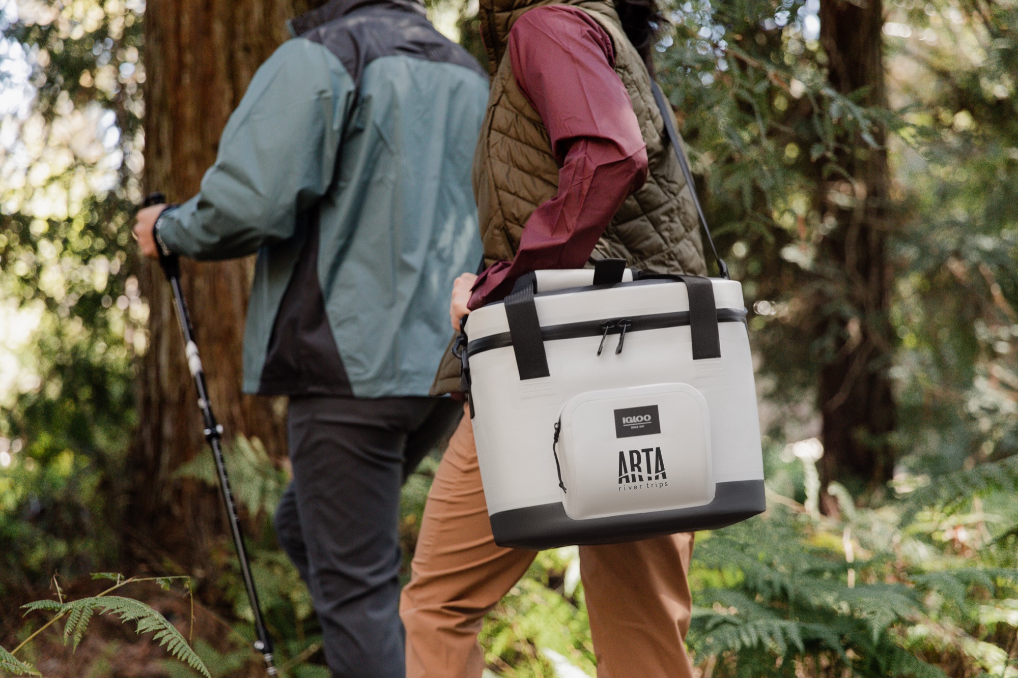 Two hikers on a forest trail carrying a soft-sided cooler with a custom printed brand decal