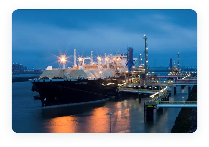 LNG carrier docked at a regasification terminal at night, with bright lights reflecting on the water and industrial infrastructure in the background.