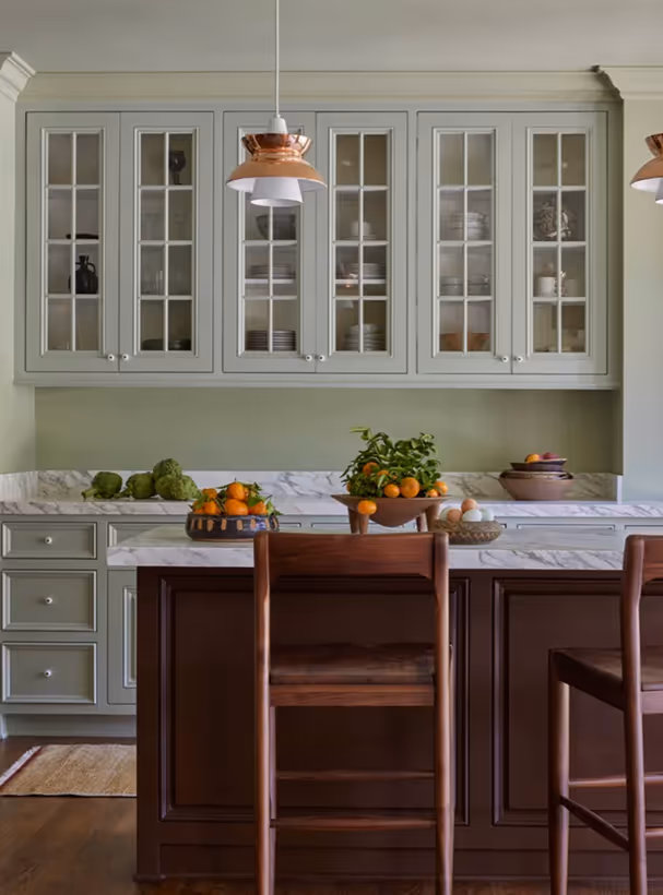 Modern kitchen with olive green cabinets, marble countertops, wooden island with chairs, and bowls of oranges and eggs on the island.