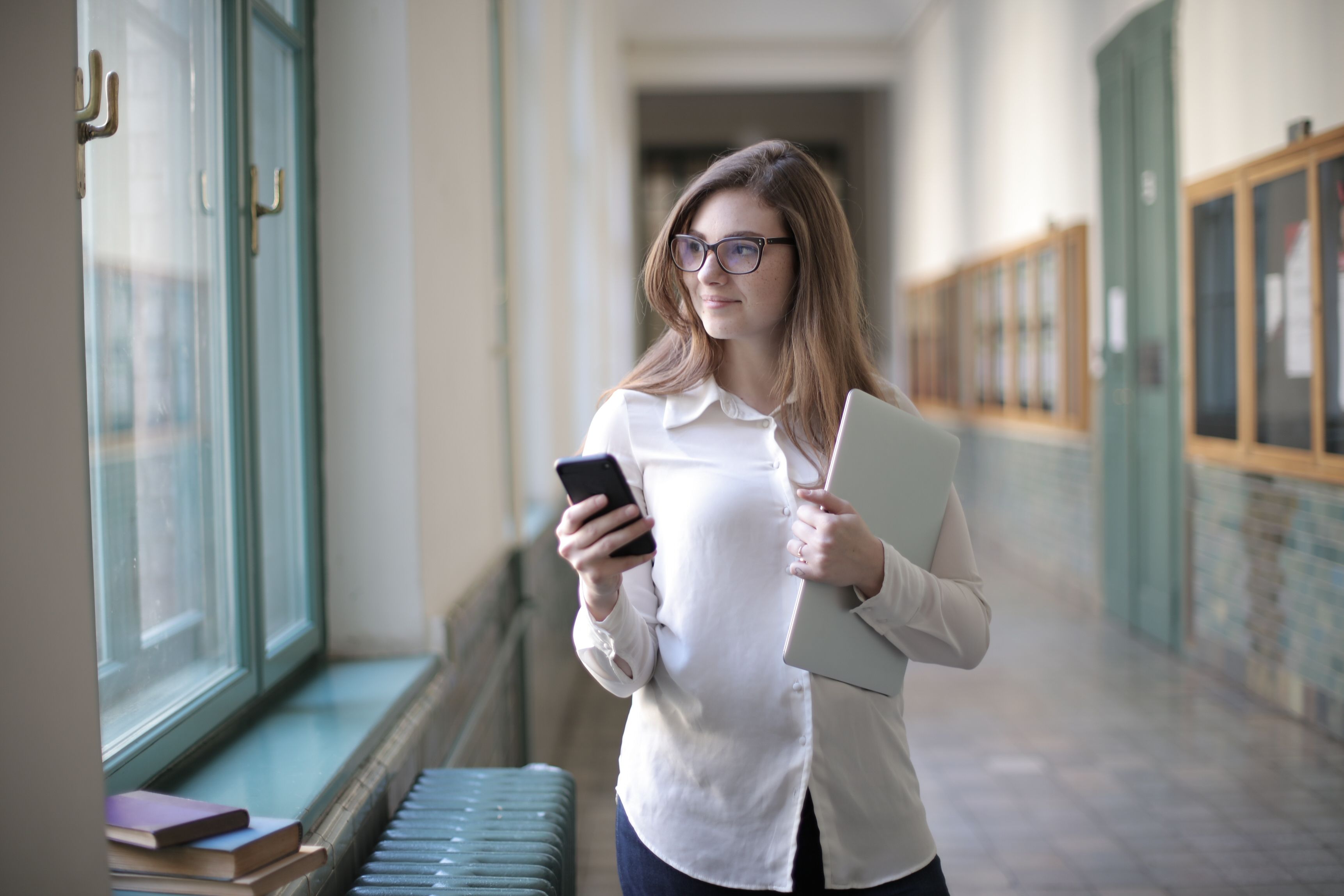 University staff member checking smartphone while carrying laptop in campus hallway