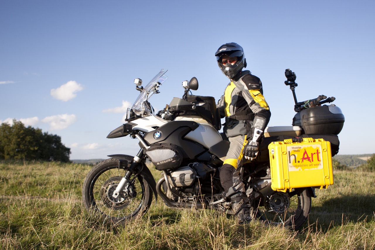 Will is gearedup and sitting on his BMW R1200 GS Adventure motorcycle standing in a field (the one above his house actually). The bike has bright yellow panniers with the h.Art logo.  on.