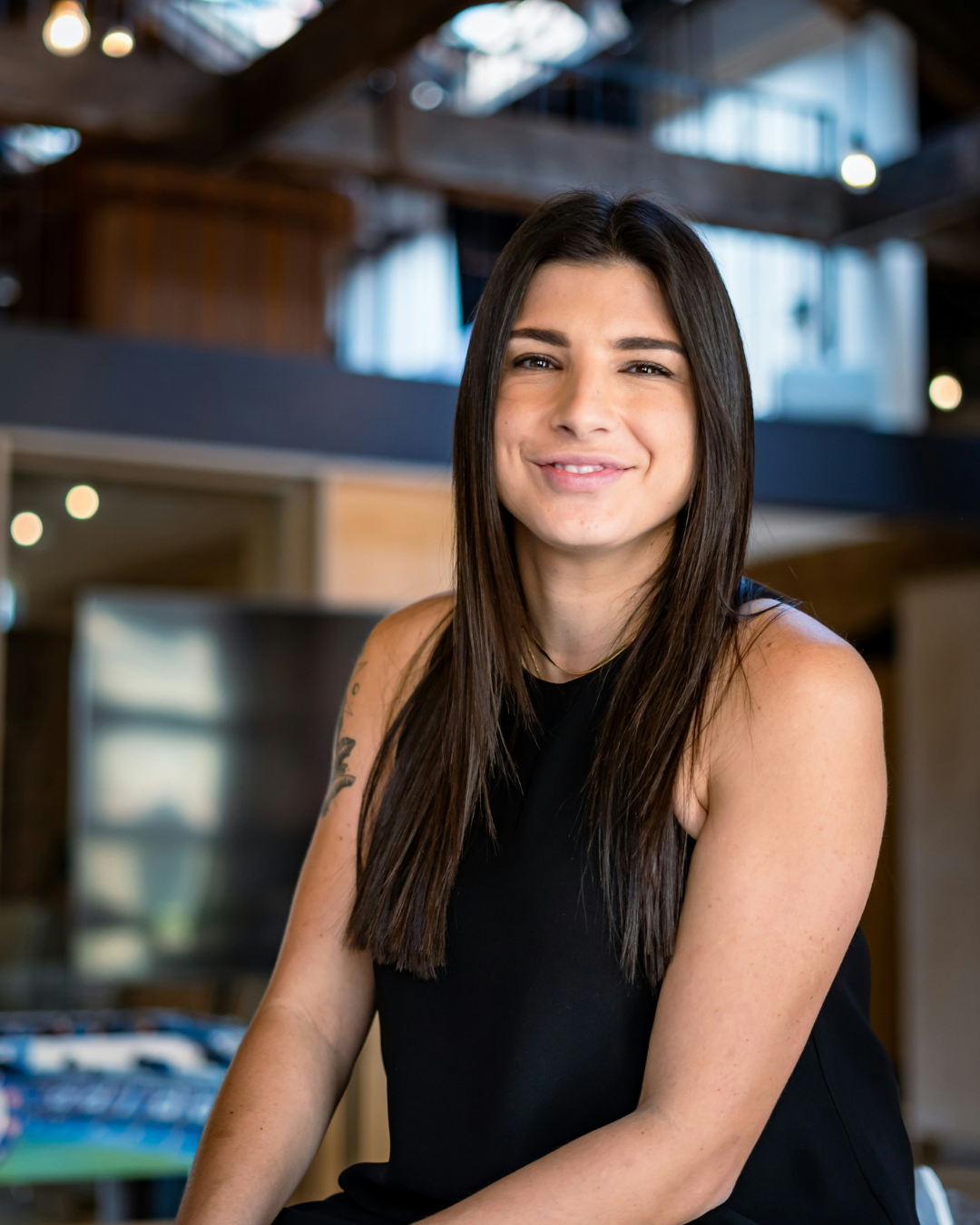 Young woman with long brown hair, wearing a white jacket and gold hoop earrings, smiling brightly outdoors with green grass in the background.