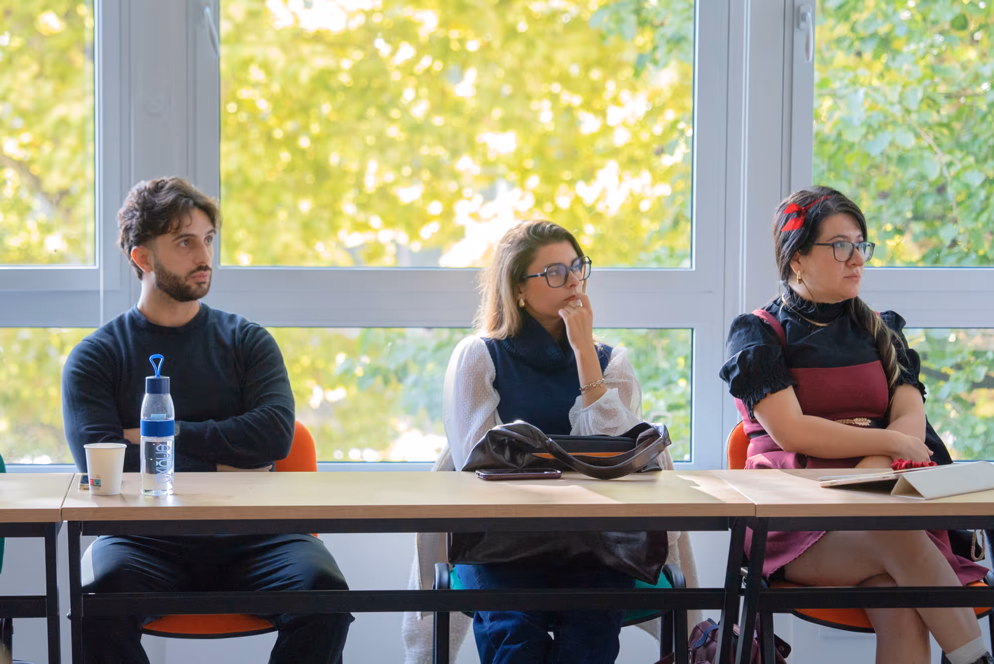 Trois adultes assis à une table devant une grande fenêtre avec vue sur des arbres verts, l'un regarde sur le côté, les deux autres semblent pensifs.