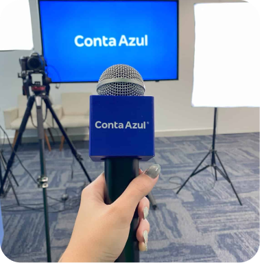 Pessoa segurando um microfone com o logo Conta Azul em um estúdio de gravação, preparado para um webinar com transmissão ao vivo, com fundo com tela digital e iluminação adequada.