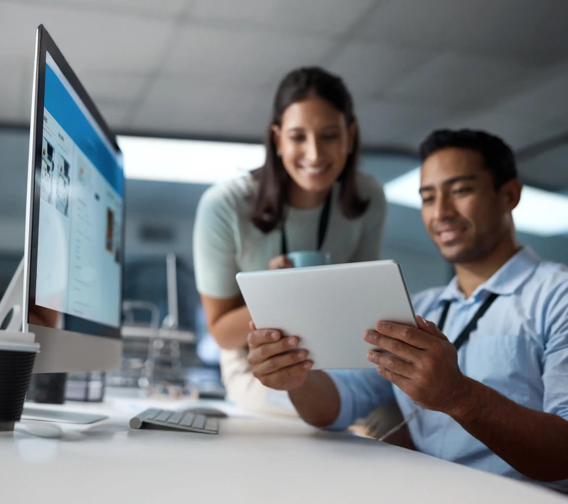 Dois colegas de trabalho sorrindo enquanto olham para um tablet em um escritório moderno com computador e acessórios na mesa.