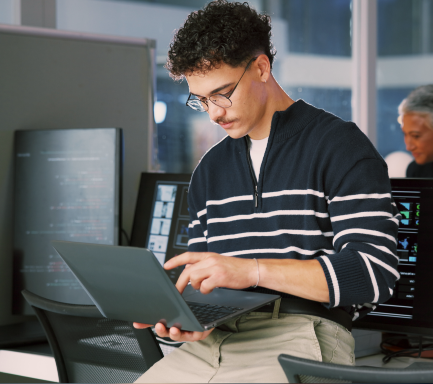 Homem com cabelo cacheado e óculos usando um laptop em um ambiente de escritório moderno com monitores ao fundo.