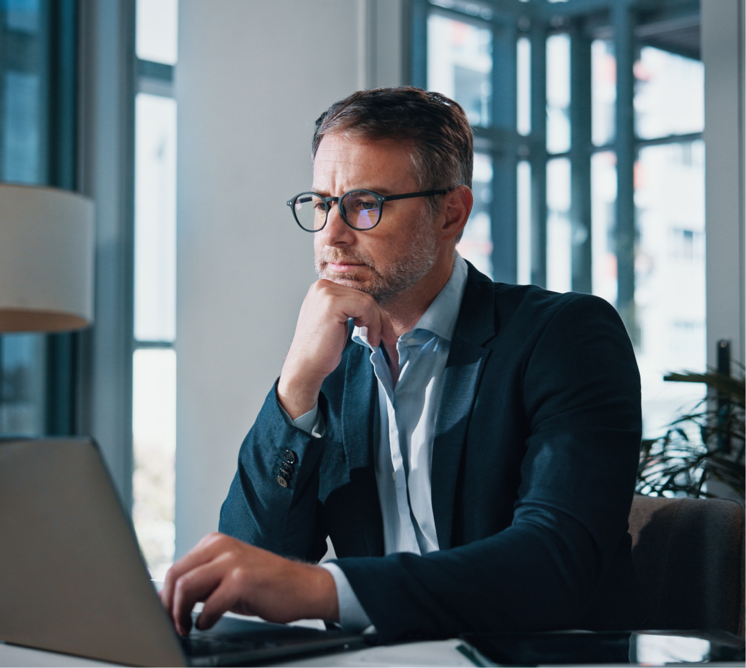 Homem de meia-idade com óculos e terno azul, pensativo, trabalhando em um laptop em um escritório moderno.