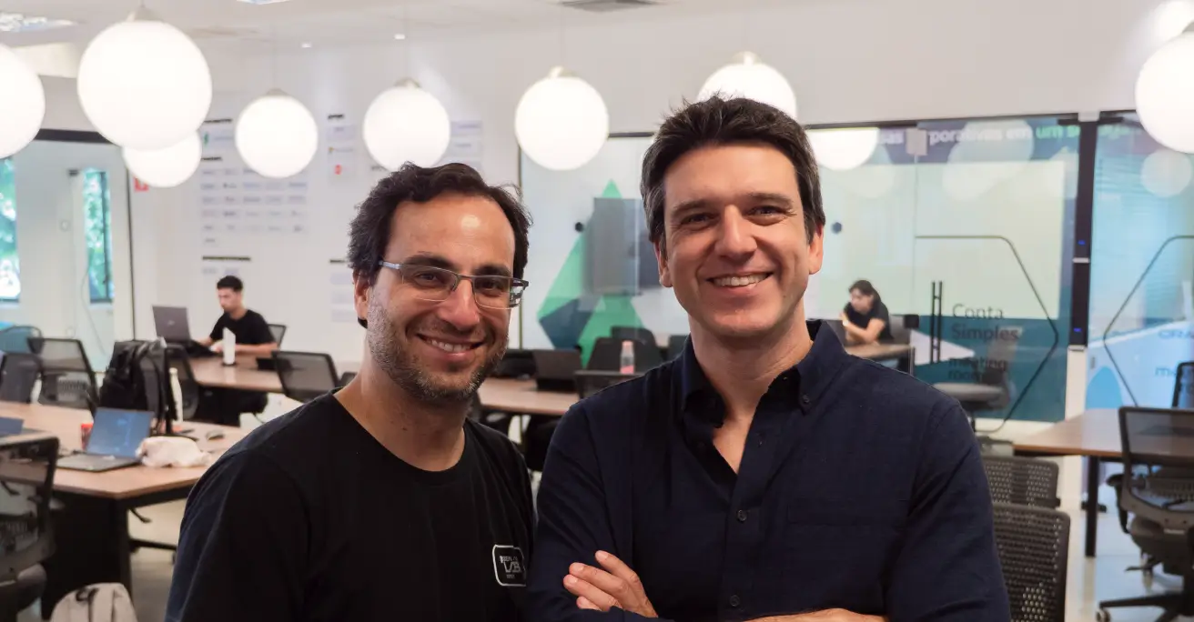 Dois homens sorridentes de braços cruzados em um escritório moderno com mesas, cadeiras e luzes pendentes.