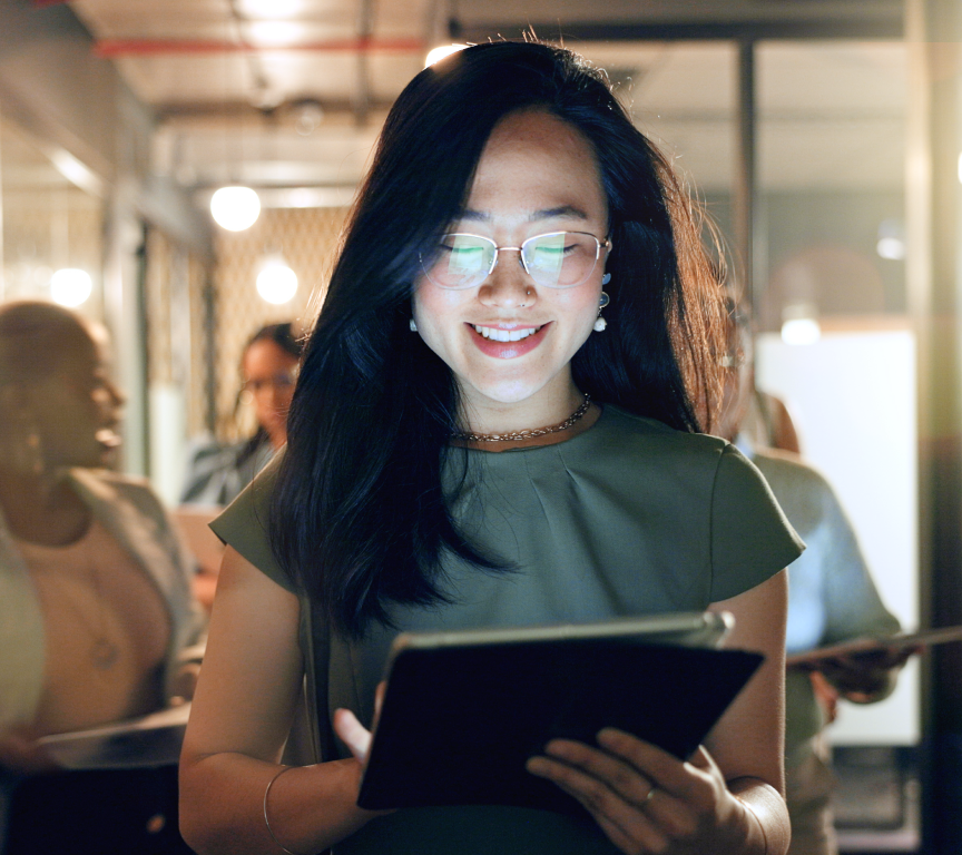 Mulher sorridente usando óculos lendo algo em um tablet em um ambiente de escritório com outras pessoas ao fundo.