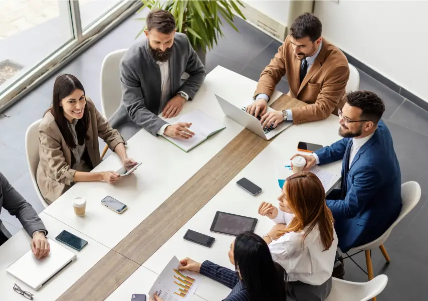 Grupo de pessoas em uma reunião de negócios ao redor de uma mesa, discutindo gráficos e usando dispositivos eletrônicos.