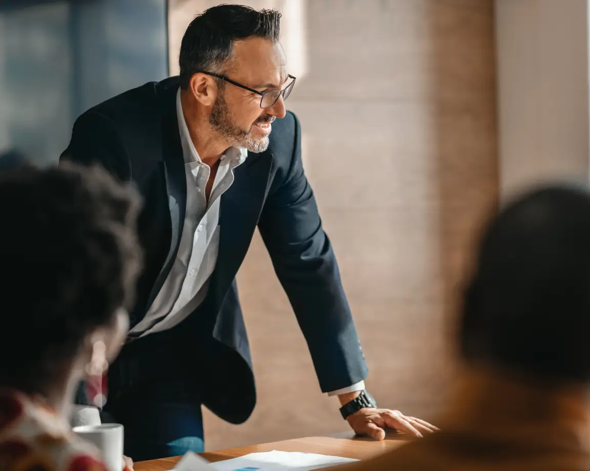 Homem de negócios com óculos e barba penteada, vestido com terno azul escuro, liderando uma reunião ao se apoiar sobre a mesa.