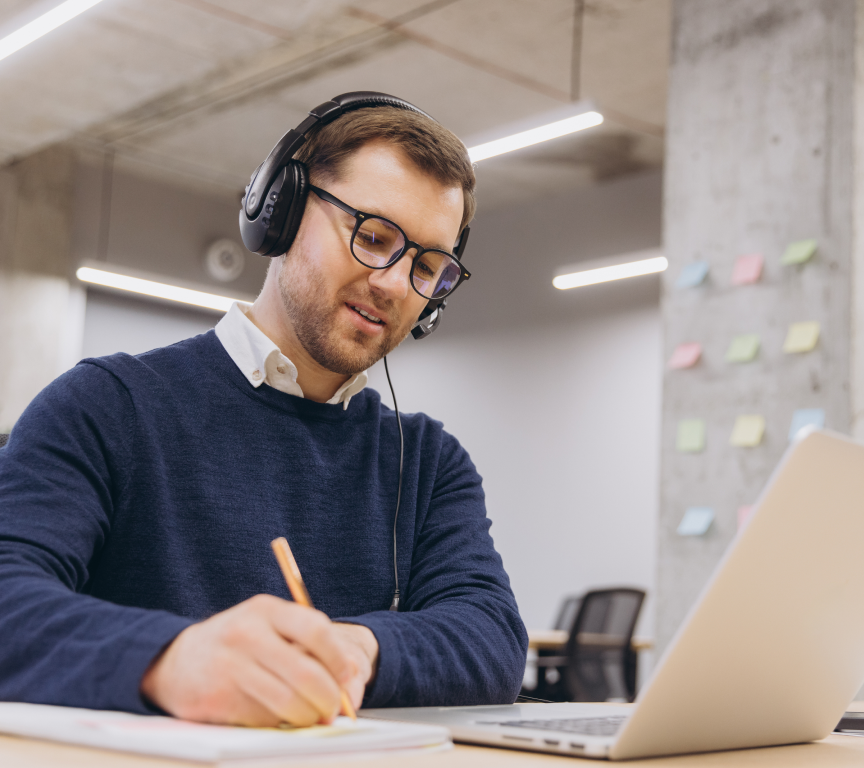 Homem usando fones com microfone e óculos escrevendo em caderno ao lado de notebook em escritório moderno.