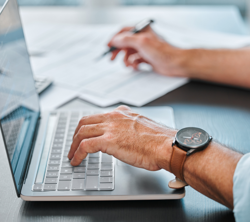 Pessoa digitando em laptop prateado com pulseira marrom enquanto escreve com caneta em documentos ao fundo.