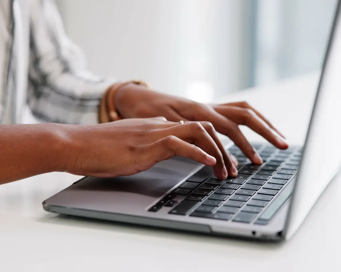 Mãos de uma pessoa negra digitando em um laptop com teclado preto.