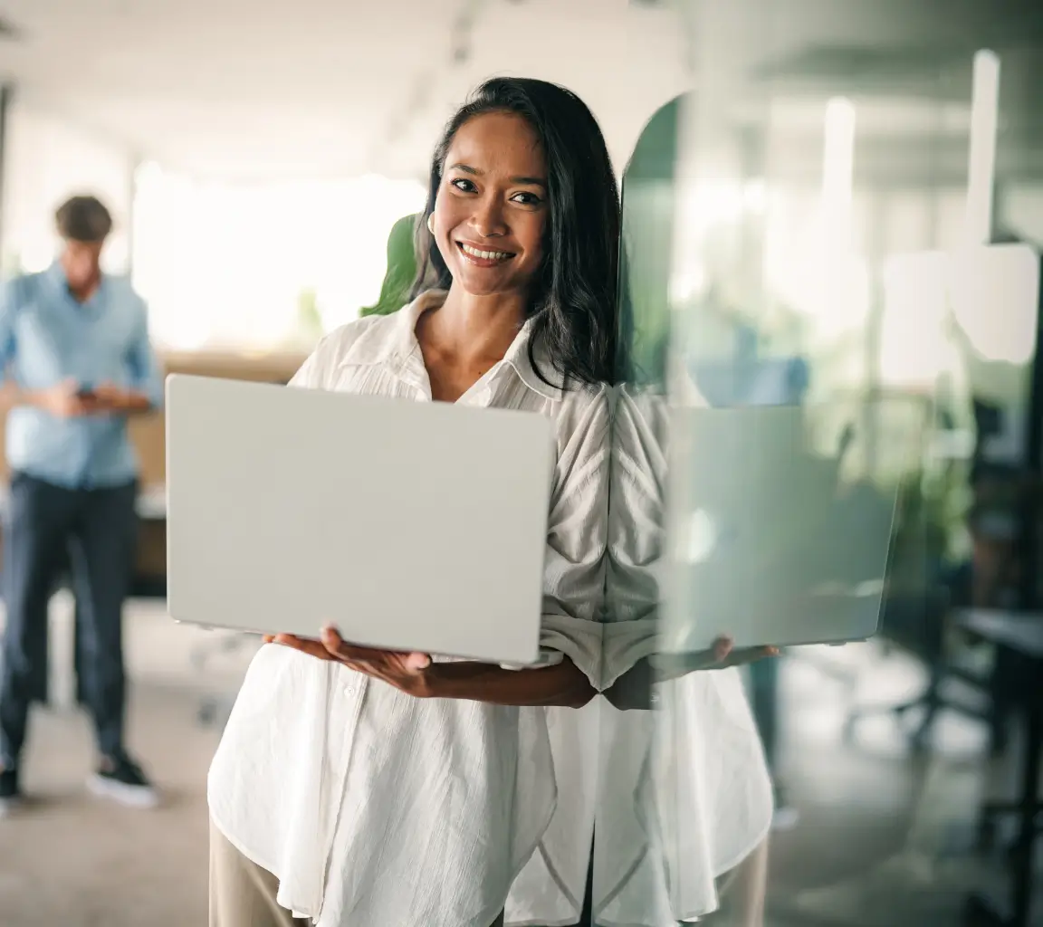 Mulher sorridente segurando um laptop em pé perto de uma parede de vidro em um escritório moderno.