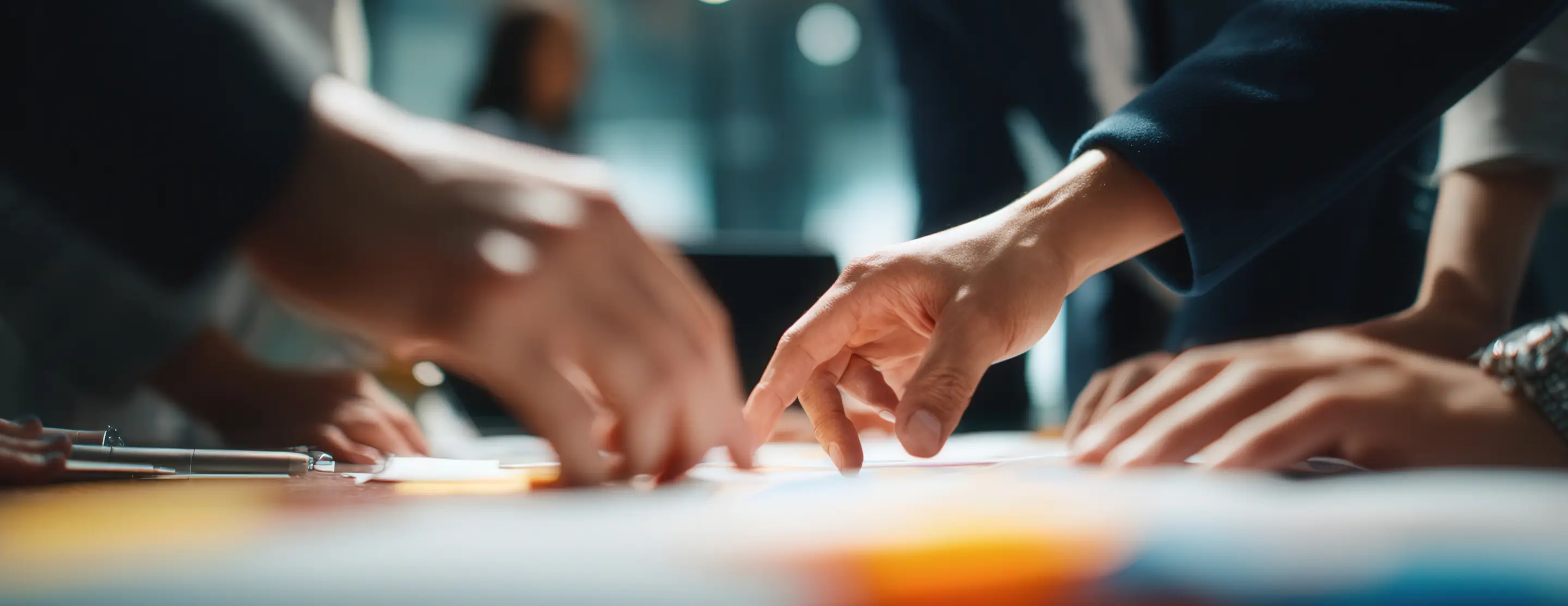Mãos de pessoas discutindo e apontando documentos sobre uma mesa em um ambiente de trabalho colaborativo.