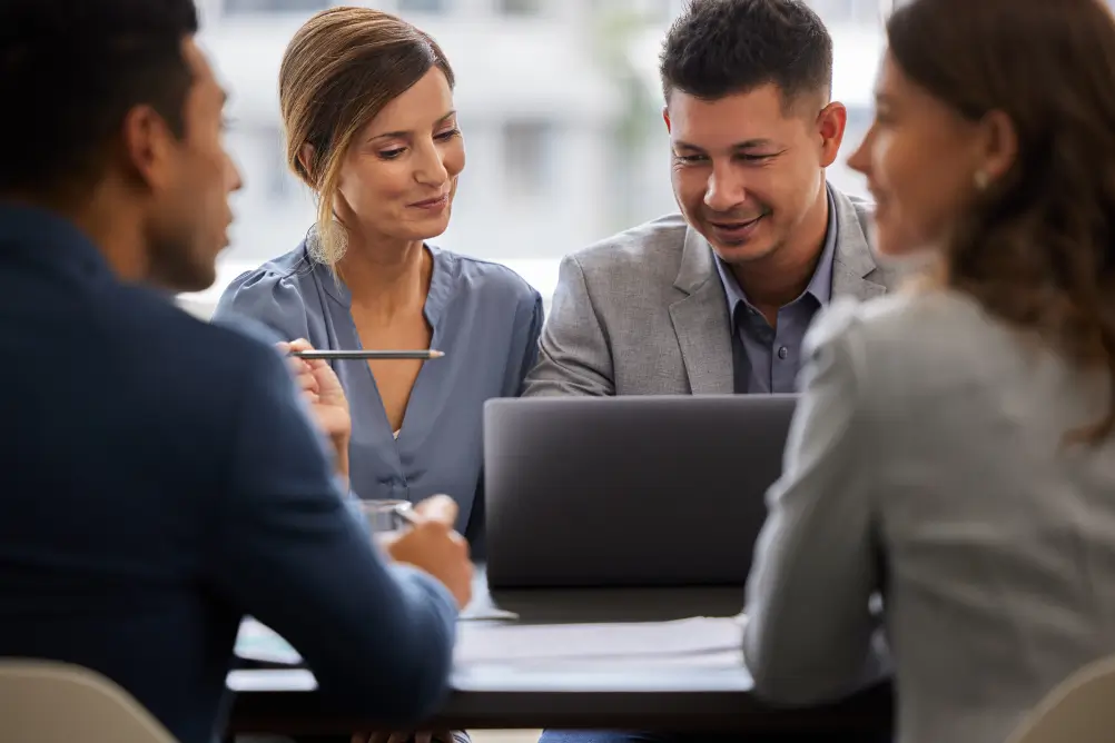 Quatro profissionais em reunião ao redor de uma mesa, olhando e discutindo conteúdos em um laptop.