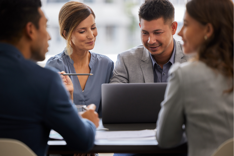 Quatro profissionais em reunião ao redor de uma mesa, olhando e discutindo conteúdos em um laptop.