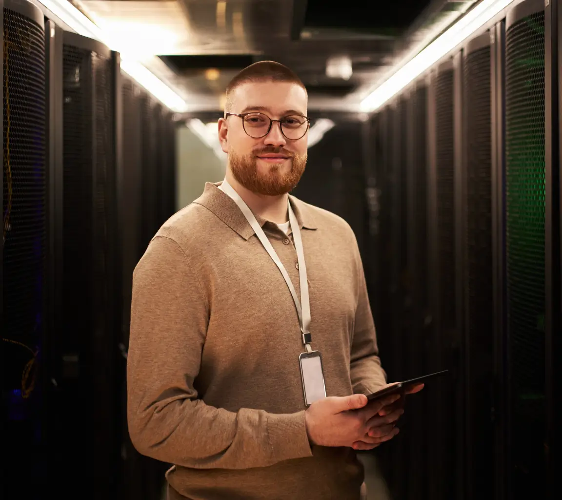 Homem com barba, usando óculos e suéter bege, segurando tablet em corredor de servidor de dados.