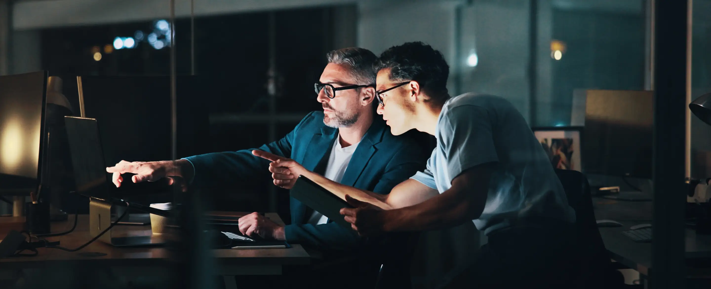 Dois homens reunidos à noite no escritório olhando para um computador e discutindo, um segurando um tablet.