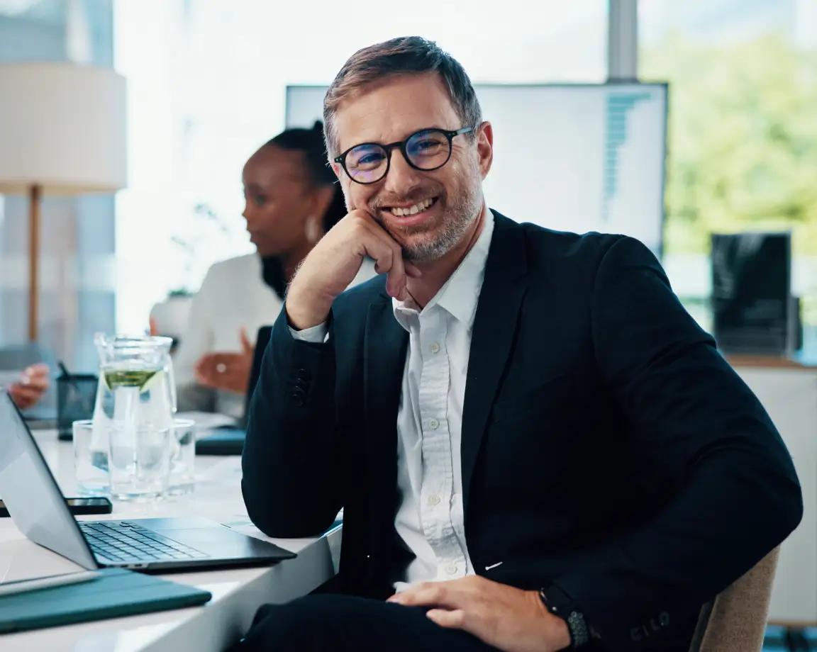 Homem sorridente de terno escuro e óculos sentado em mesa de escritório com notebook aberto, com colegas de trabalho ao fundo.
