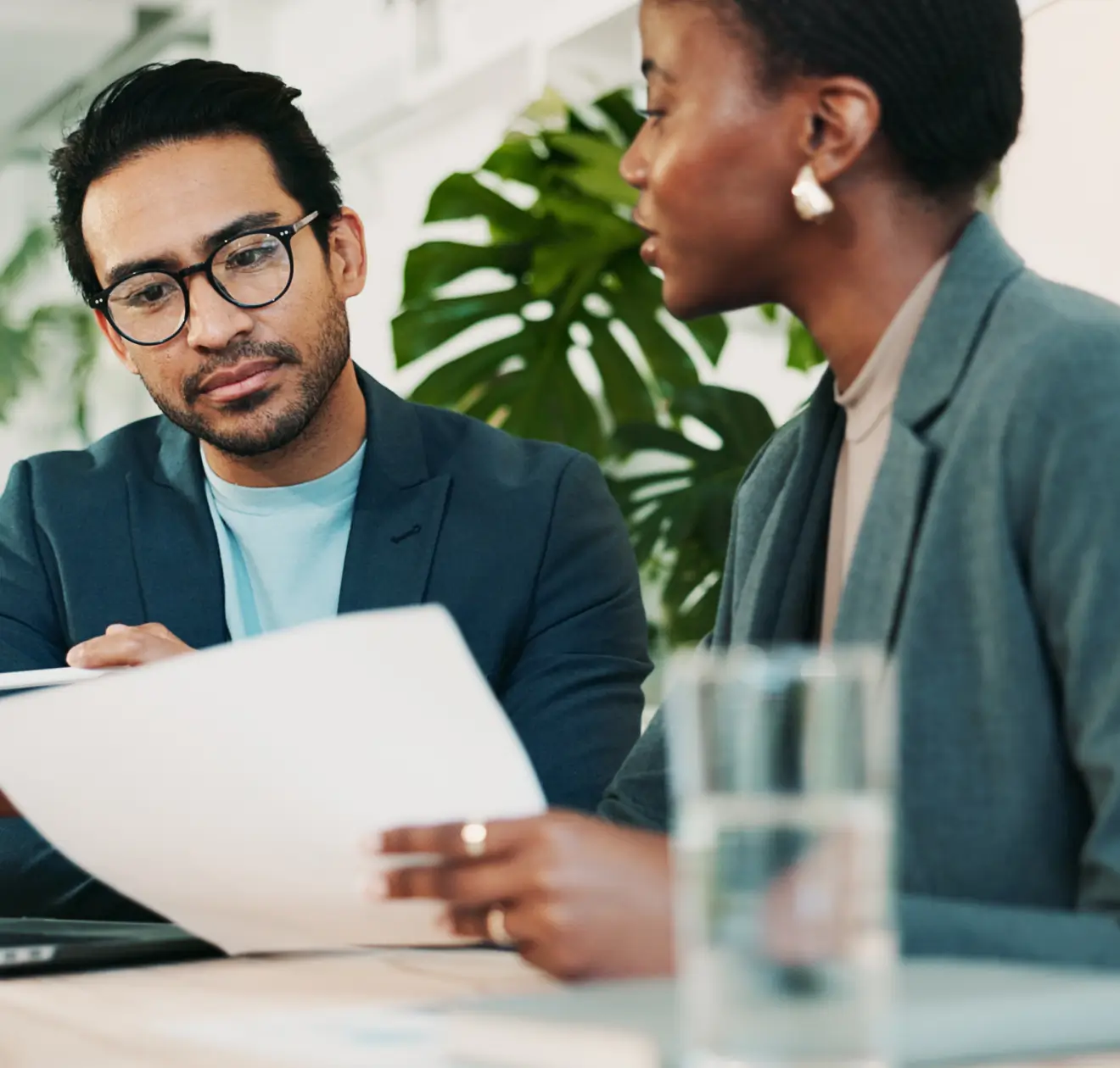Dois profissionais vestidos de maneira formal discutindo documentos em um escritório com plantas ao fundo.