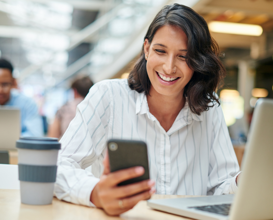 Mulher sorridente olhando para o celular em uma mesa com laptop e copo reutilizável em um ambiente de trabalho moderno.