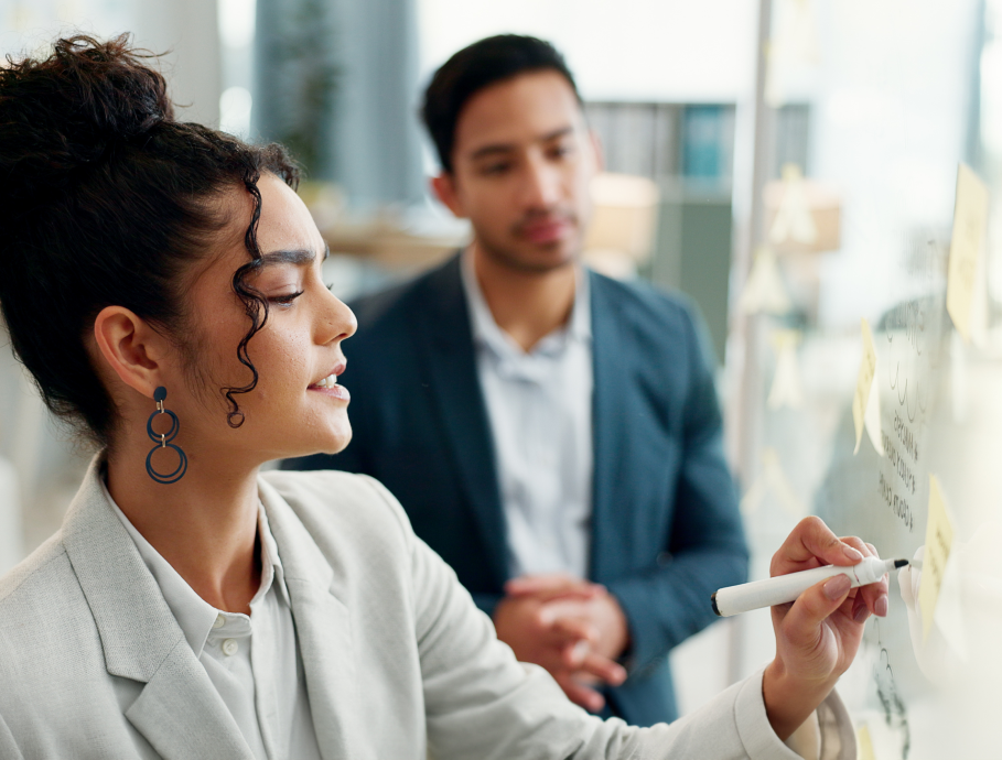 Mulher escrevendo em um quadro branco com post-its, enquanto um homem a observa em um ambiente de escritório.