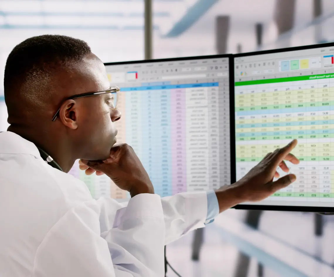 Homem em bata branca olhando e apontando dados em duas telas de computador exibindo planilhas coloridas.