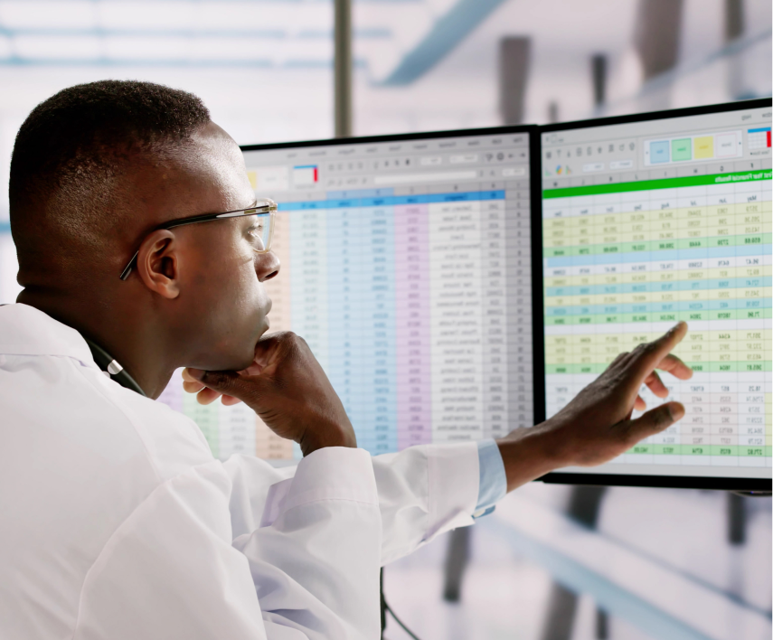 Homem em bata branca olhando e apontando dados em duas telas de computador exibindo planilhas coloridas.