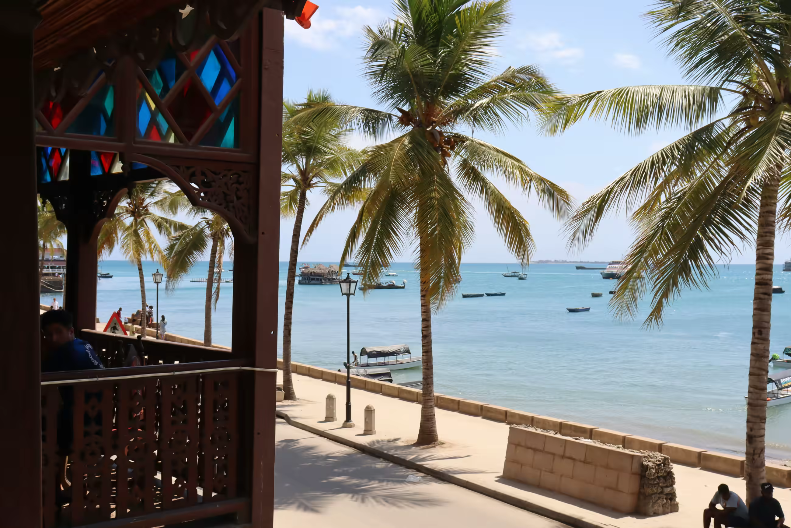 Palm trees line a seaside promenade with boats on calm blue water under a clear sky.
