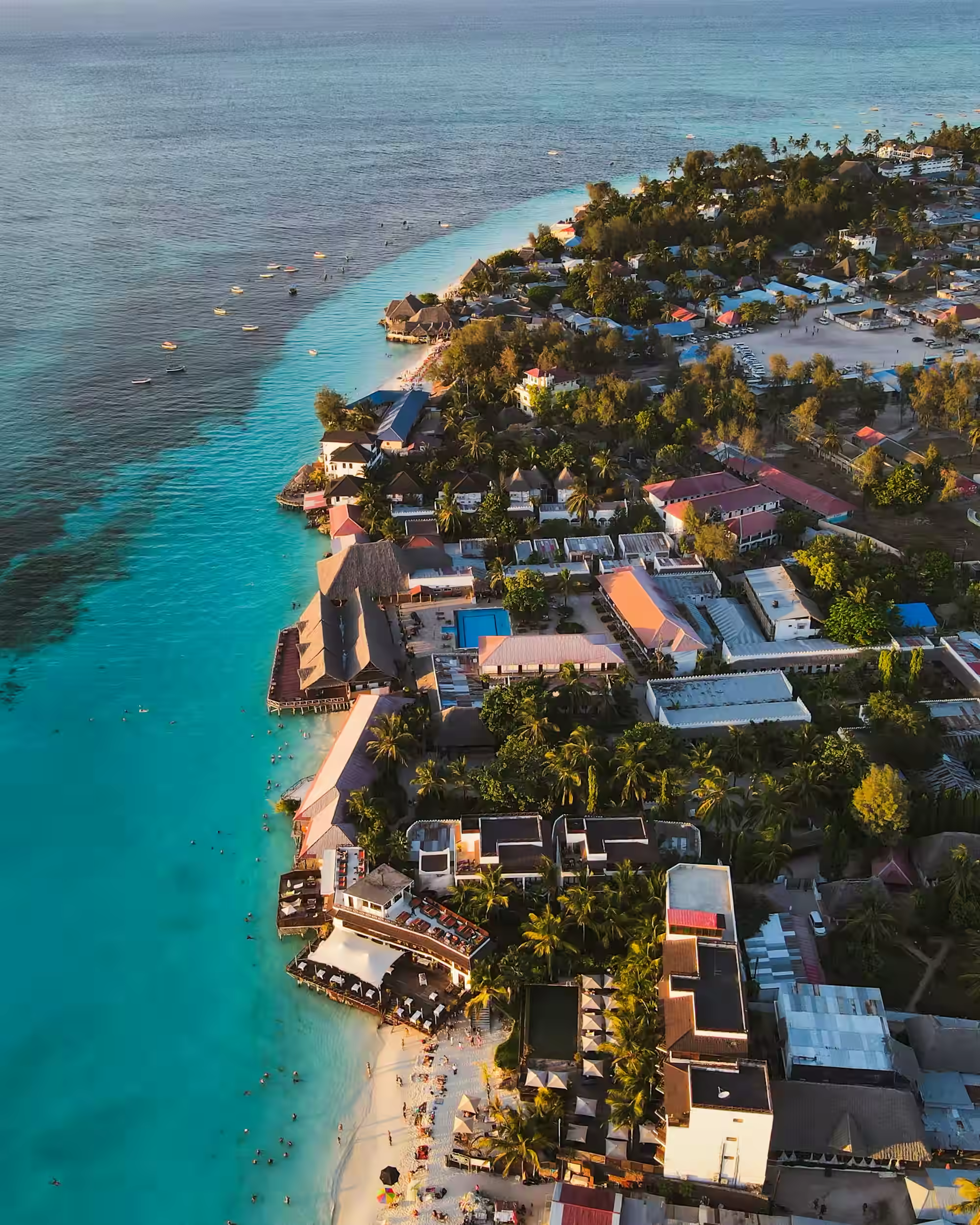 Aerial view of the Zanzibar coastline at golden hour, with turquoise shallows, a reef edge and palm-shaded resorts along a white sand beach
