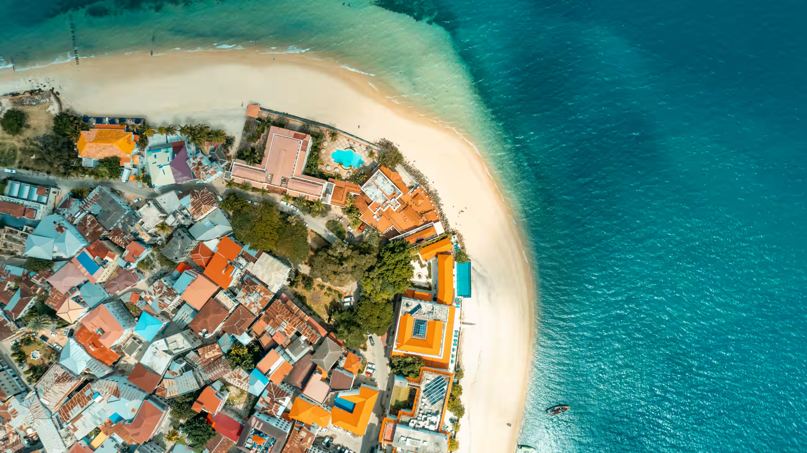 Overhead view of Stone Town's terracotta rooftops meeting the shoreline, with a curving sand beach and deep blue water