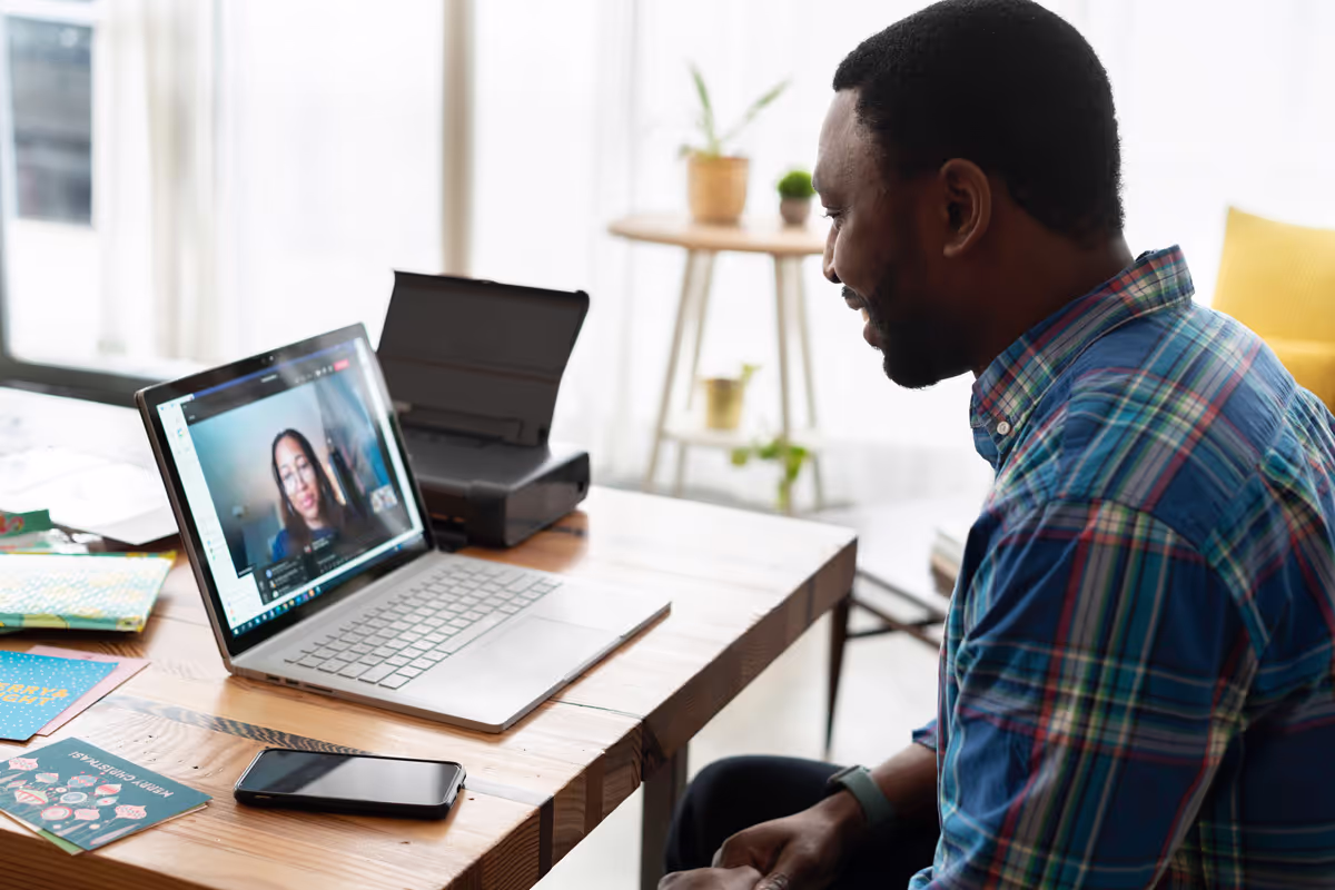 Man in a blue plaid shirt video chatting with a woman on a laptop at a wooden desk.