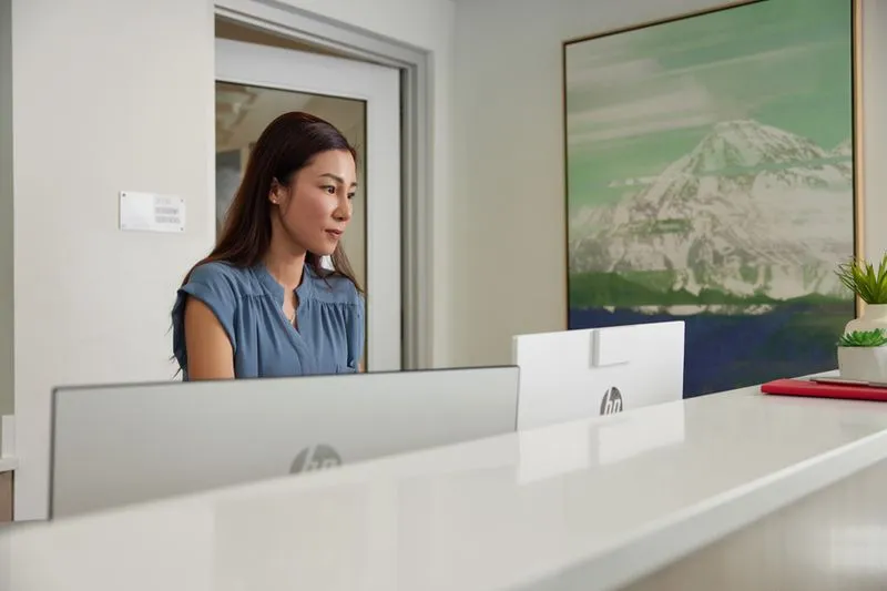 Woman with long dark hair in blue blouse working at a reception desk with two HP monitors.