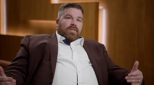 Bearded man in a white shirt and dark blazer speaking with expressive hand gestures against a blurred wooden background.