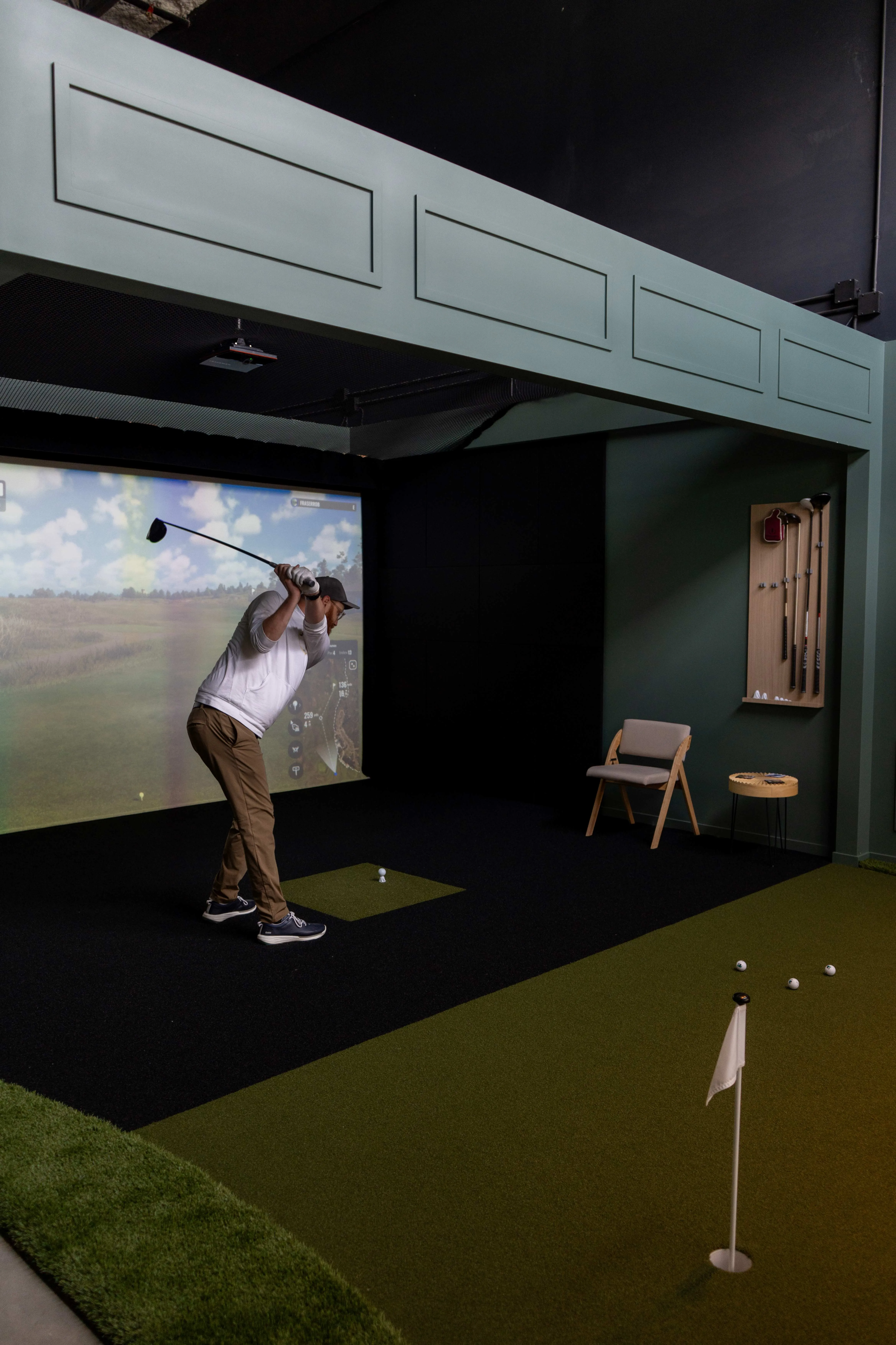 Man swinging golf club in front of indoor golf simulator screen with putting green and golf balls inside room.