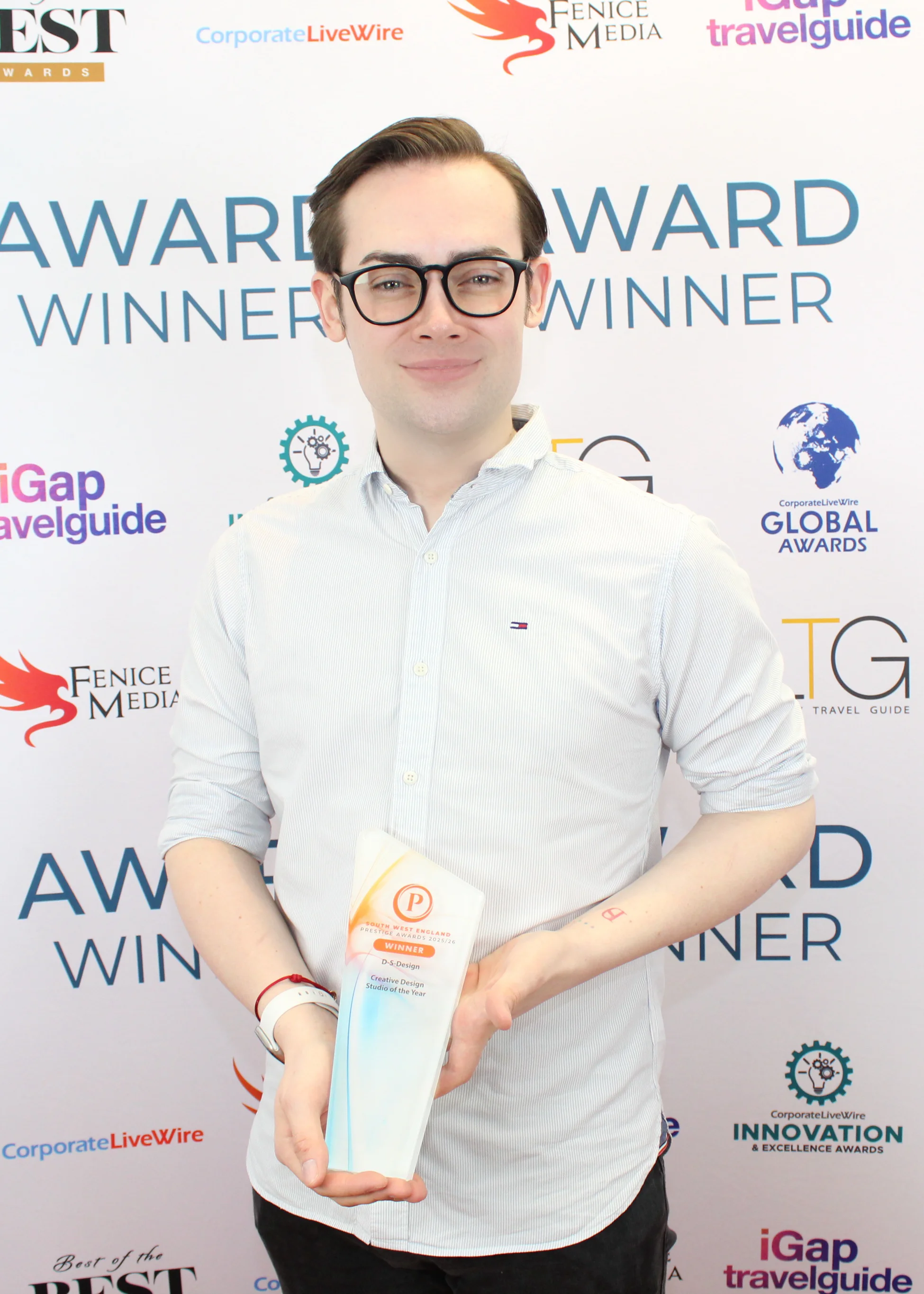 A young man wearing glasses and a light striped shirt smiles while holding a South West England Prestige Awards 2025/26 trophy for Creative Design Studio of the Year, standing in front of a backdrop with award and sponsor logos.