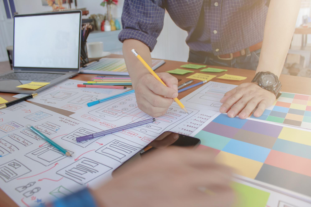 A designer sketching website wireframes beside a laptop displaying a completed web design mockup.