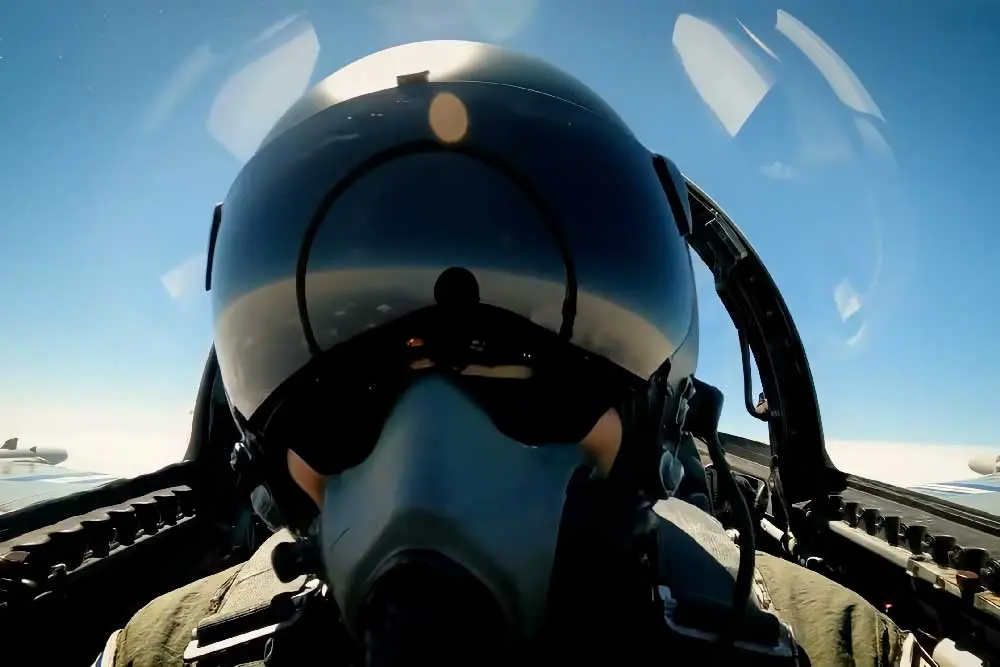 ine Nahaufnahme eines Kampfjet-Piloten im Cockpit, der einen Helm und eine Sauerstoffmaske trägt. Der Himmel ist klar und blau.