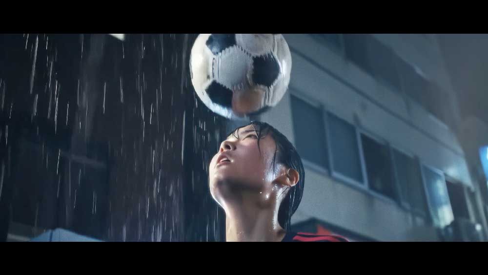 Eine junge Fussballspielerin jongliert nachts im Regen einen Fussball mit dem Kopf. Ihre Haare sind nass und der Fussball ist in der Luft über ihr zu sehen.