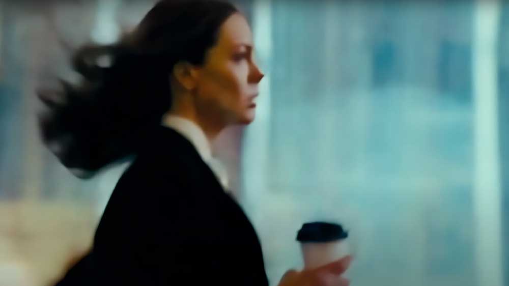 Eine Frau im Profil eilt mit einem Kaffeebecher in der Hand an einem großen Fenster vorbei. Ihre Haare wehen im Wind.