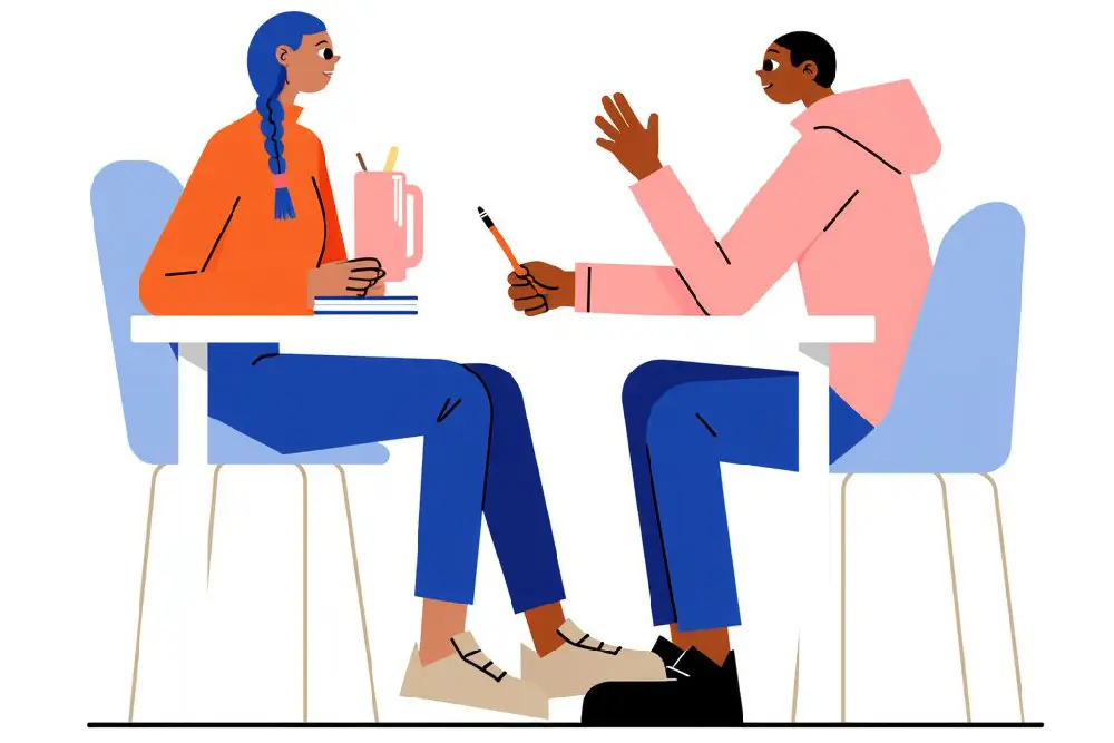 Zwei Personen sitzen sich an einem Tisch gegenüber und unterhalten sich. Die eine Person mit blauen Zöpfen und einem orangefarbenen Pullover sitzt vor einer Kaffeetasse und Notizbüchern, die andere Person mit einem pinken Kapuzenpullover gestikuliert mit den Händen.