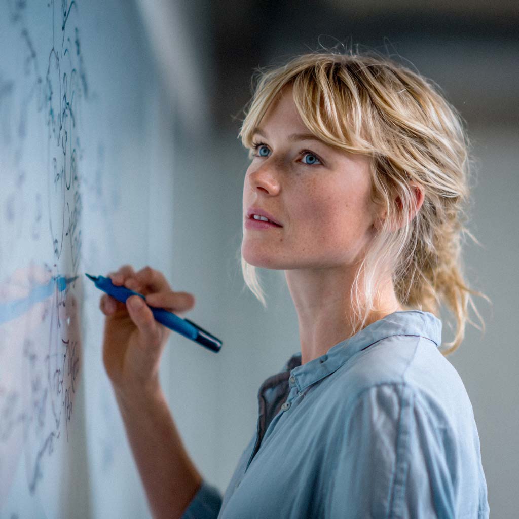Eine Frau mit blonden Haaren und einem hellblauen Hemd schreibt mit einem blauen Stift auf ein Whiteboard. Sie blickt nachdenklich nach oben und hat Sommersprossen im Gesicht.