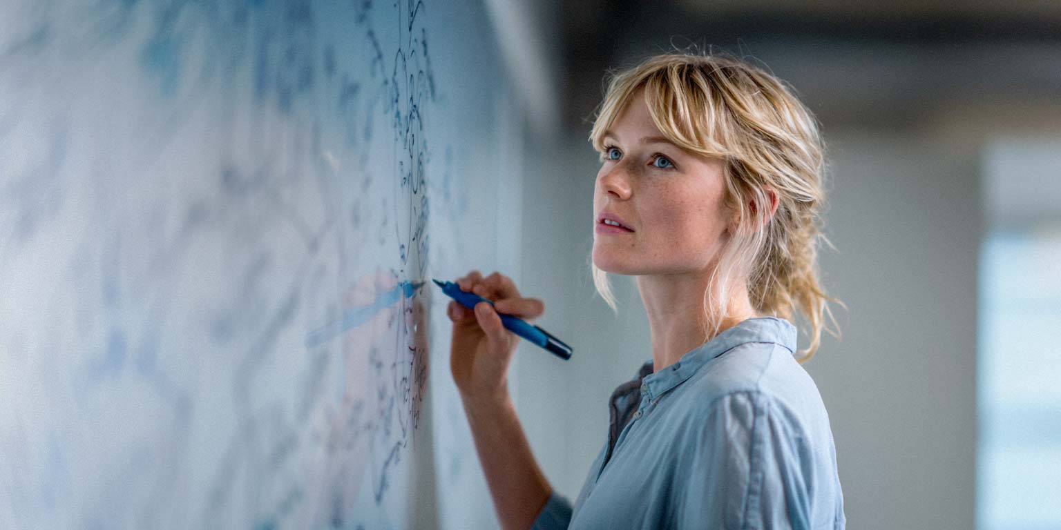 Eine Frau mit blonden Haaren und einem hellblauen Hemd schreibt mit einem blauen Stift auf ein Whiteboard. Sie blickt nachdenklich nach oben und hat Sommersprossen im Gesicht.