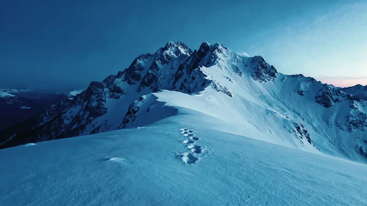 Eine tiefblaue, dämmerungsartige Aufnahme einer schneebedeckten Berglandschaft. Im Vordergrund führt eine geschwungene Schneewehe über einen Grat, in der deutliche Fussspuren zu sehen sind. Dahinter erheben sich mehrere schroffe, von Schnee bedeckte Gipfel, die im blauen Licht dramatisch wirken.