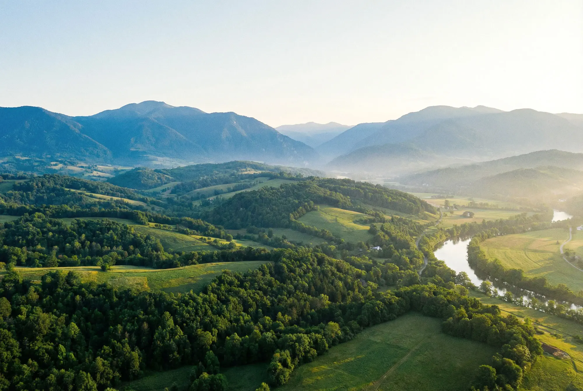 Luftaufnahme von grünen, bewaldeten Hügeln und einem Fluss vor einer blauen Bergkette.