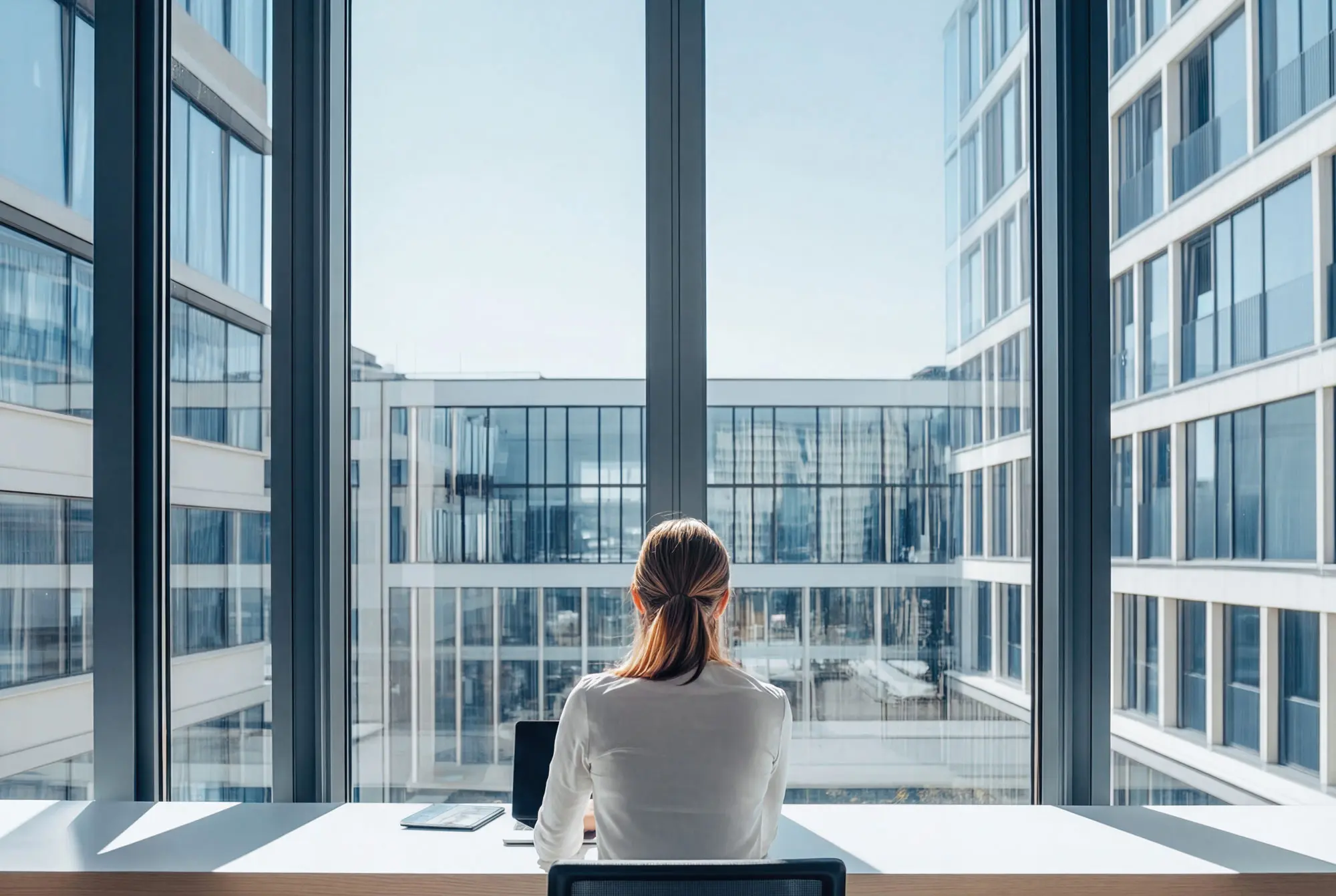 Frau am Schreibtisch im Büro, blickt auf den Laptop vor einer Glasfassade.